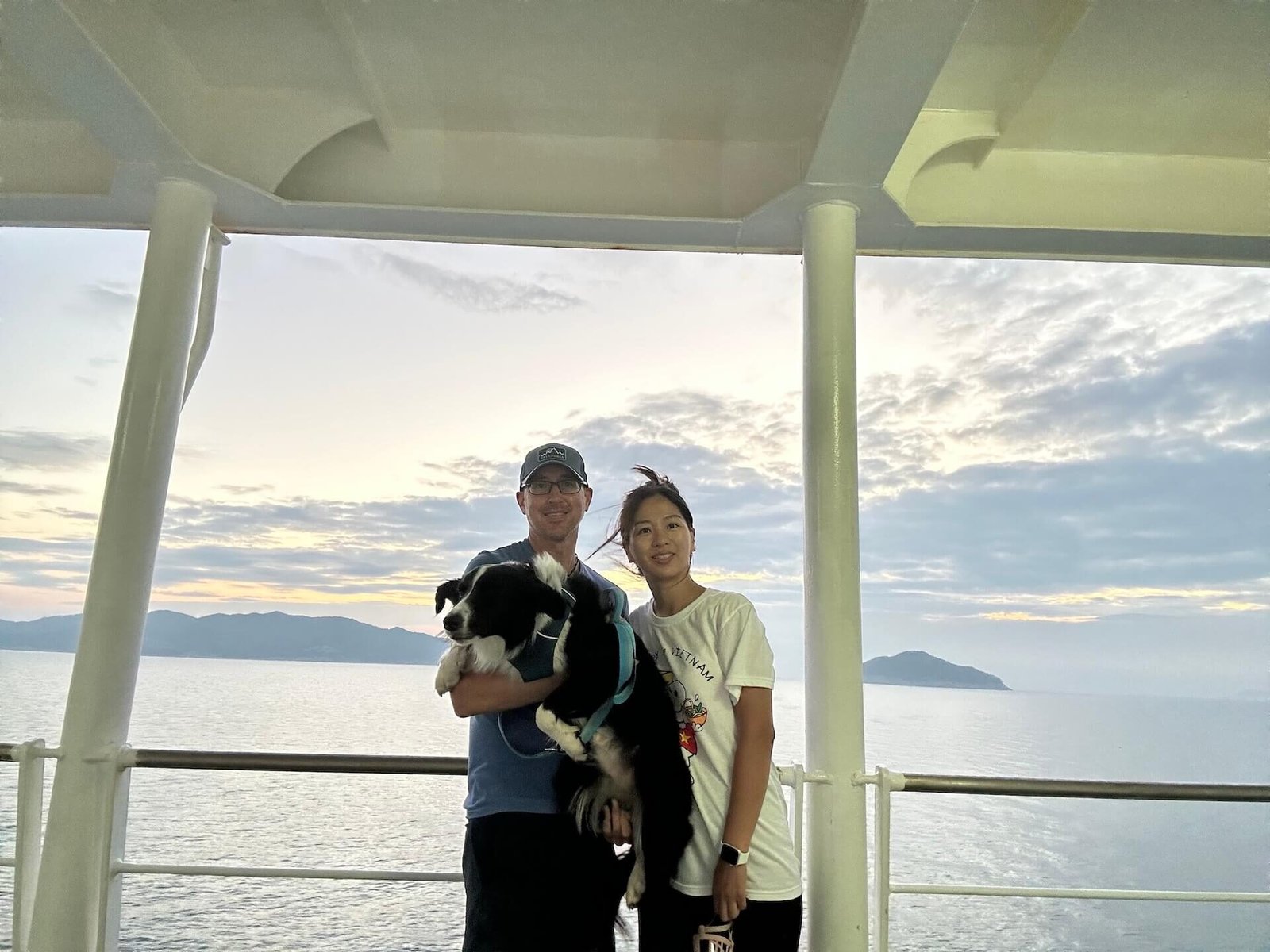 A couple with their black and white dog on a ferry to Jeju Island, South Korea, with the ocean and mountains in the background at sunset.