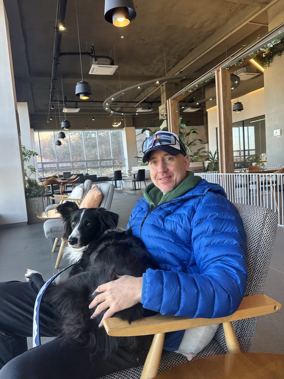 A man in a blue jacket sitting in a cozy chair at the Pure Sheep Ranch Café in Pyeongchang, South Korea, holding his border collie on his lap. The café features modern decor, large windows, and a warm atmosphere.