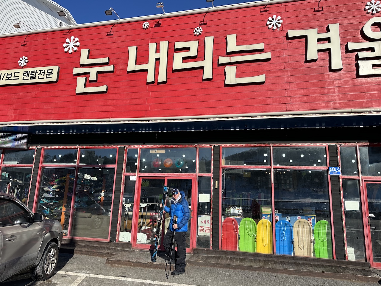 A person is standing outside a ski rental shop in Pyeongchang, South Korea, holding a newly rented pair of skis. The rental shop is one of many on the road leading to Mona Yongpyong Resort.