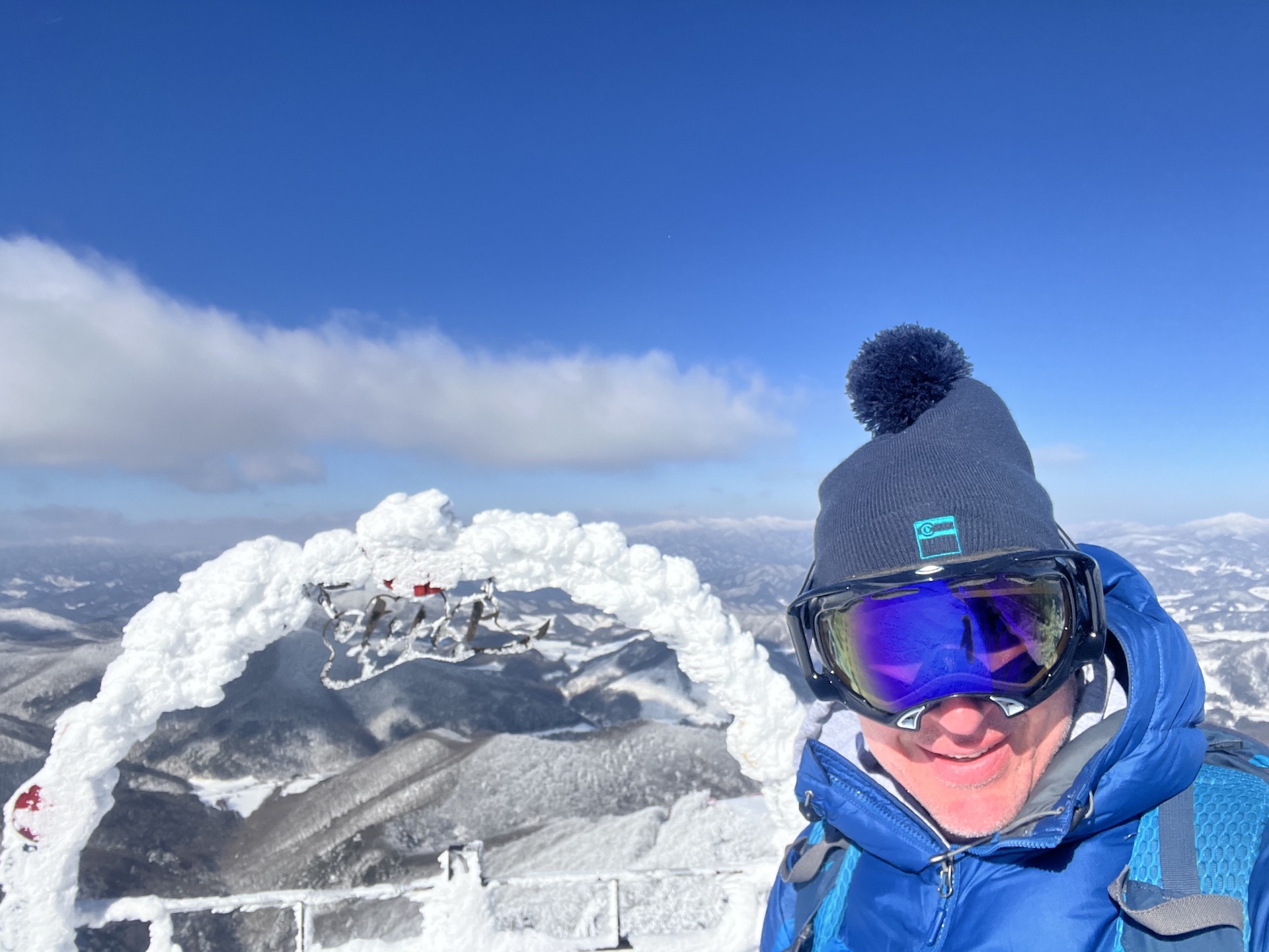 Skier standing at a Mona Park viewpoint at Yongpyong Resort with snow-covered mountains and a clear blue sky in the distance during winter.