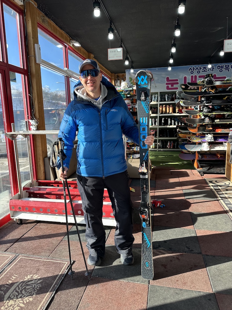 Skier standing in a rental shop near Pyeongchang, holding rental skis and poles.