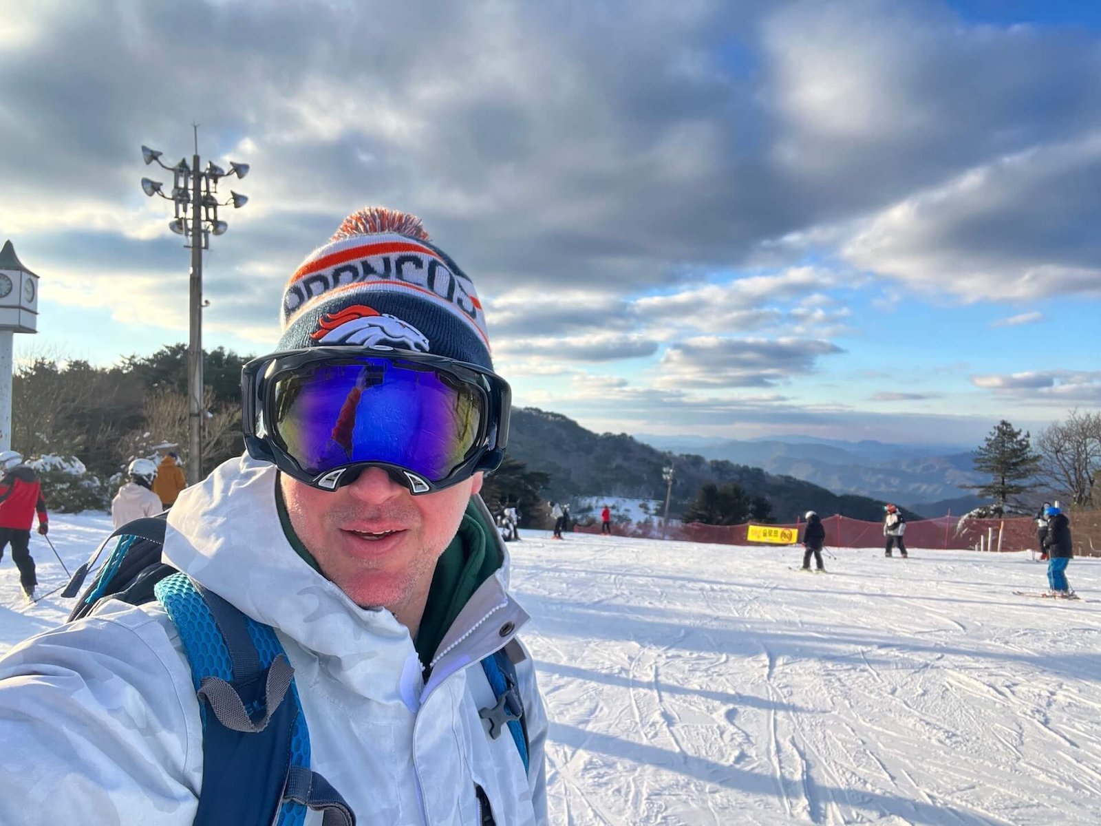 A skier in a Denver Broncos beanie at the top of the Liner Lift at Muju Deogyusan Resort in South Korea, with a view of snow-covered slopes, other skiers, and a scenic mountain backdrop under a partly cloudy sky.