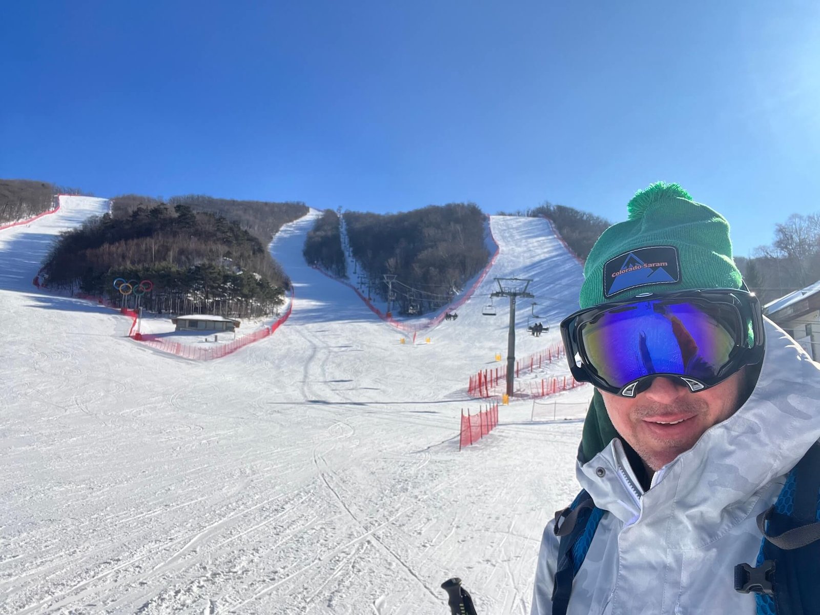 A skier wearing a green Colorado Saram beanie and goggles stands at the base of the Rainbow Zone at Yongpyong Resort, South Korea, with Olympic slalom courses and a chairlift visible in the background on a sunny winter day.