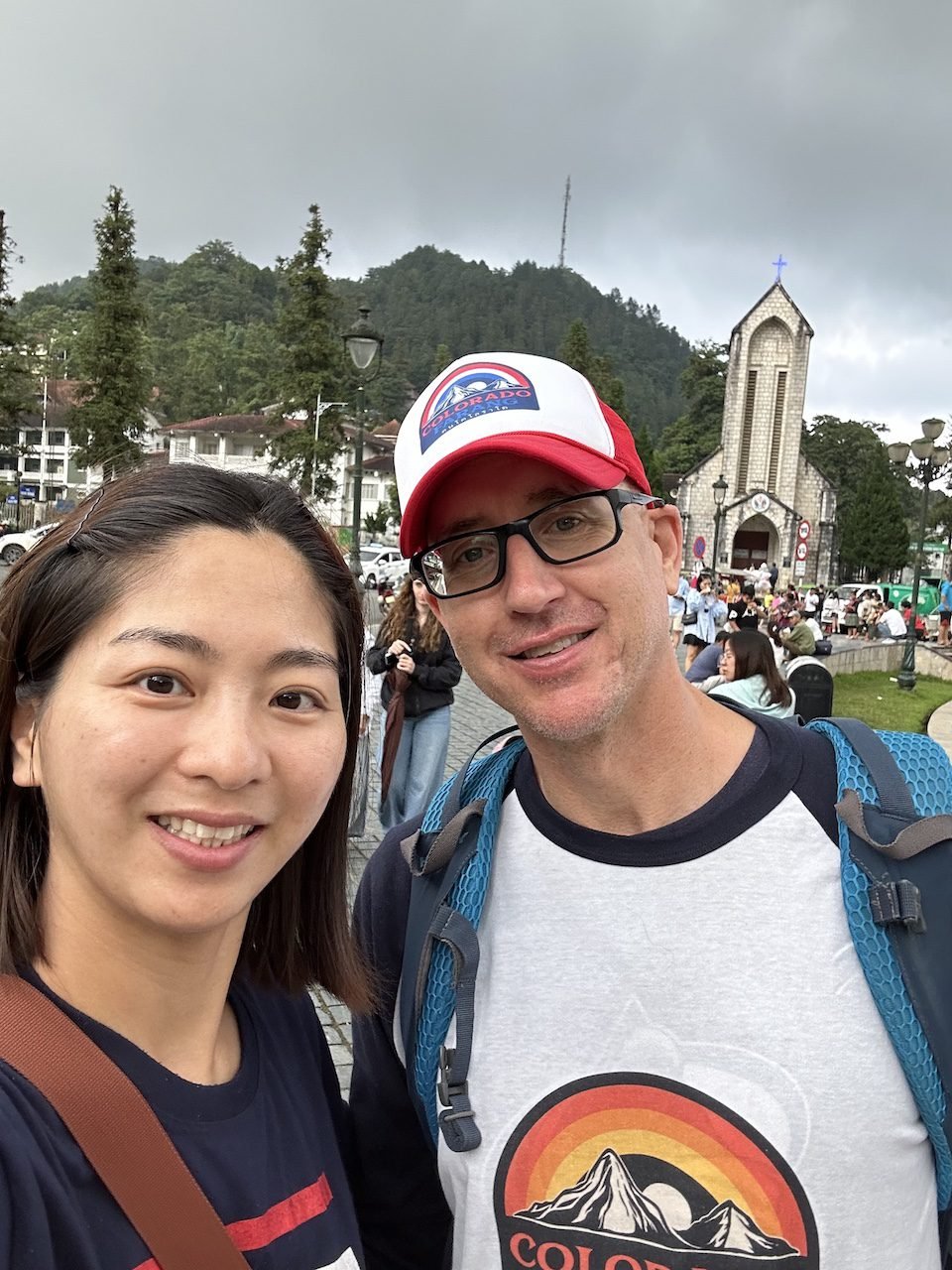 A couple smiling in Sa Pa Town Square, located across from Sun Plaza. The area features a historic stone church, green hills, and a lively atmosphere with visitors enjoying the open space.