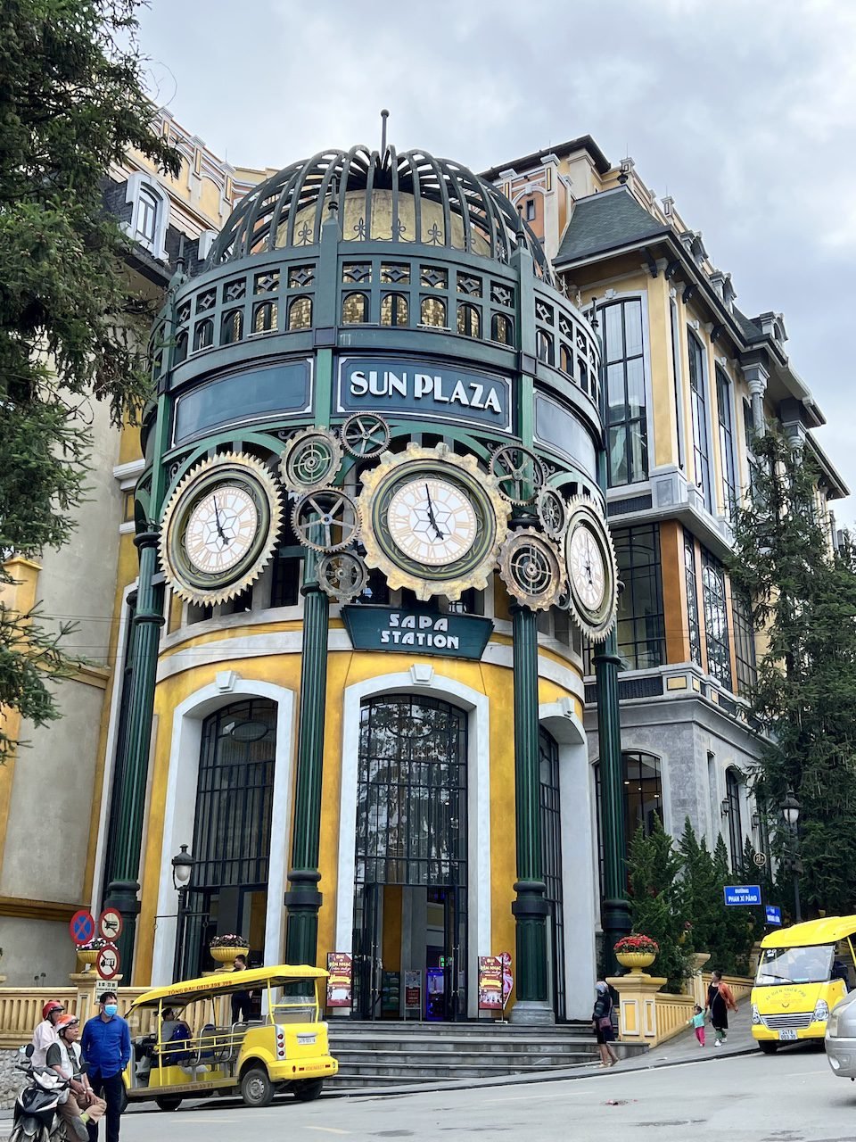 The Sun Plaza building in Sa Pa Town, Vietnam, featuring a bright yellow facade, large clock tower, and entrance to Sapa Station, surrounded by transport vehicles and visitors.