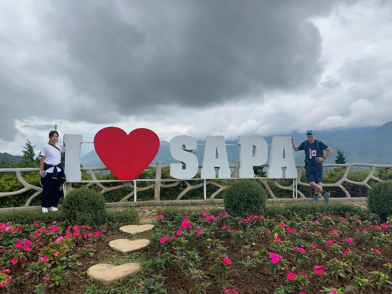 "I Love Sapa" sign surrounded by vibrant flowers, with two travelers posing at the base of the Fanispan cable car station in Sapa, Vietnam, against a backdrop of cloudy skies and lush mountain scenery.