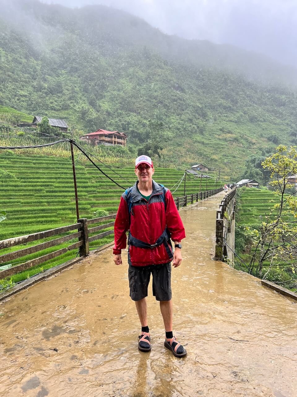 A man in a red rain jacket and black shorts stands on a muddy path in Sa Pa, Vietnam, during a rainy season trek, with misty green terraced hills in the background.