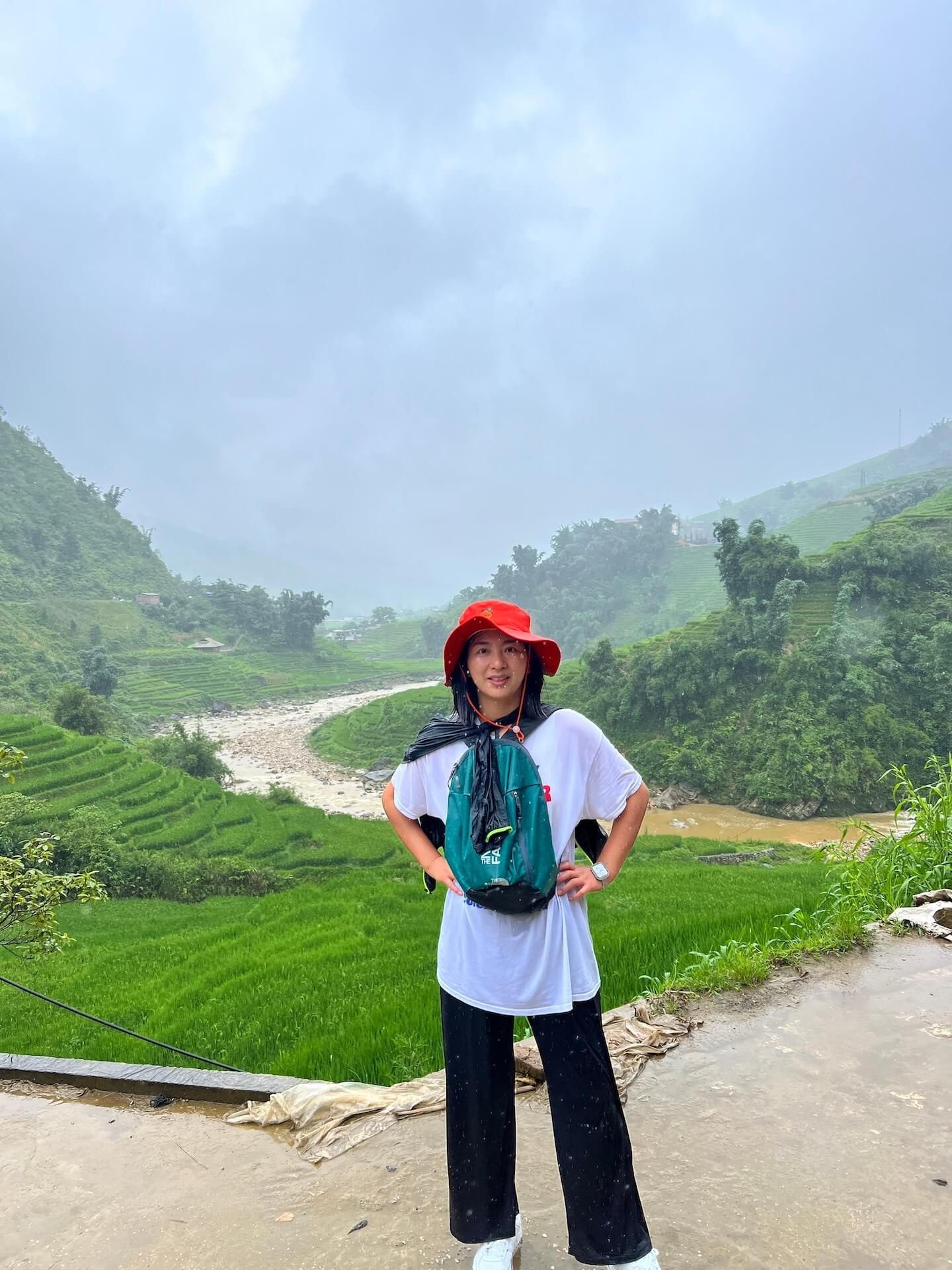 A woman standing in the rain during a self-guided trek in Sa Pa, Vietnam, surrounded by lush green rice terraces and a winding river.