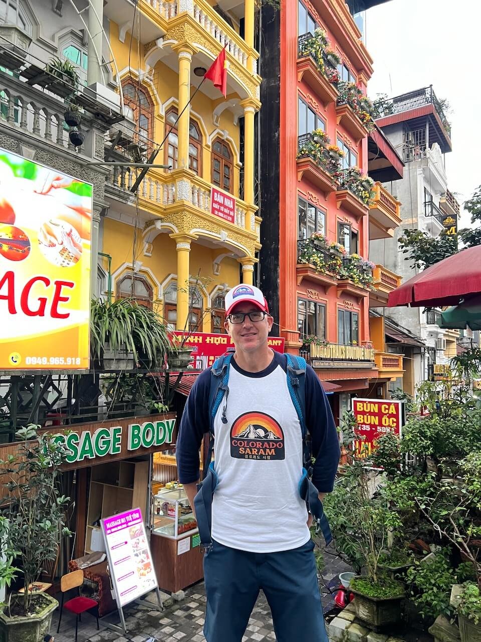 Traveler wearing a Colorado Saram t-shirt posing on a backstreet in Sapa, Vietnam, with colorful French-inspired buildings and local businesses in the background.