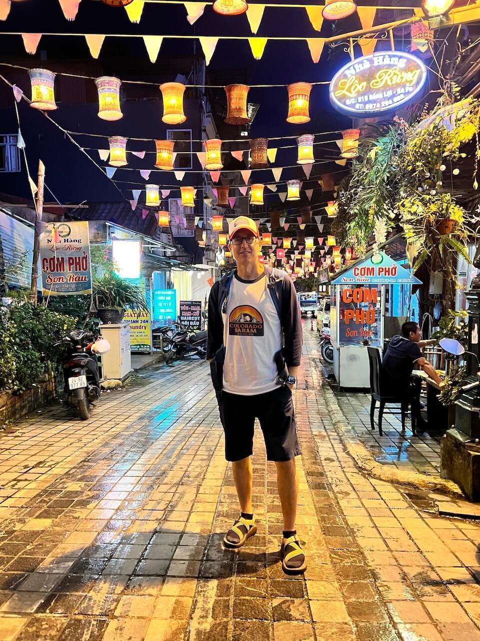 Man wearing a Colorado Saram t-shirt posing under decorative lanterns at a night market in Sapa, Vietnam, with illuminated signs, street vendors, and a lively atmosphere.