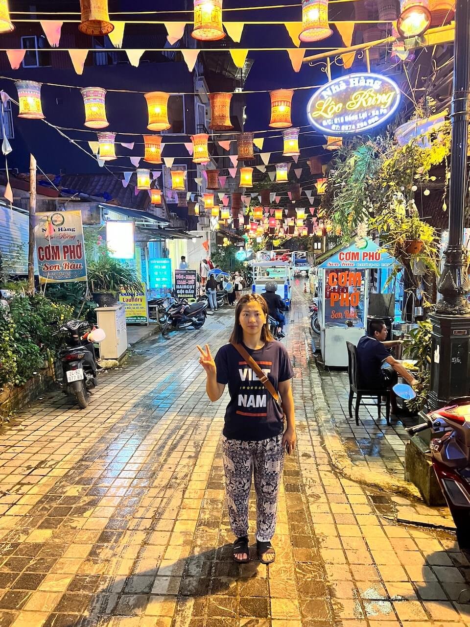 Woman wearing a "Vietnam" t-shirt posing under colorful lanterns at a night market in Sapa, Vietnam, with bustling street vendors and motorbikes in the background.