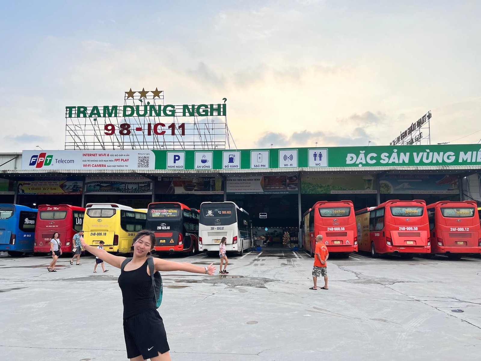  A row of colorful sleeper buses parked at a rest stop between Hanoi and Sapa, with a woman posing in front wearing a black outfit and backpack.