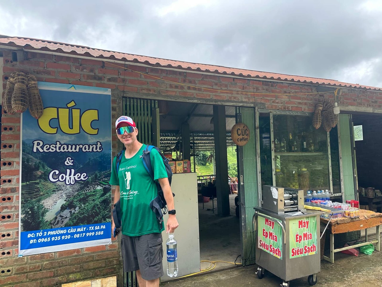 A traveler in a green hiking shirt stands in front of Cúc Restaurant & Coffee, a small eatery along the trekking route from Sapa to Lao Chai, Vietnam. The rustic building features a sign and a fresh sugarcane juice cart.