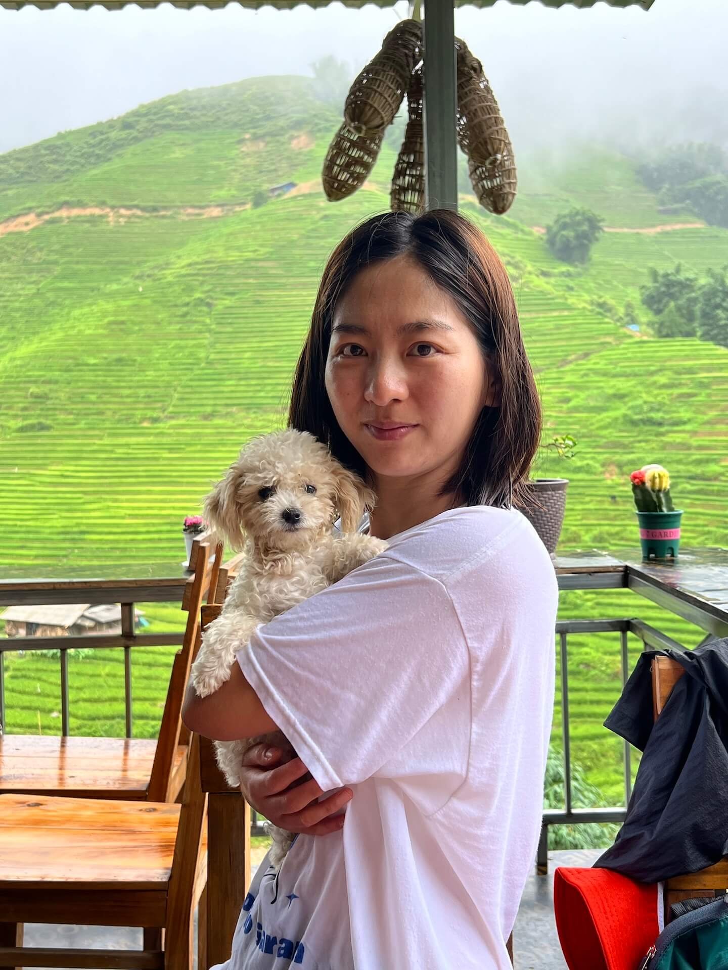 A woman holds a small fluffy dog at Cúc Restaurant & Coffee, a small eatery along the trekking route from Sapa to Lao Chai. Behind her, lush green rice terraces stretch across the misty hills.