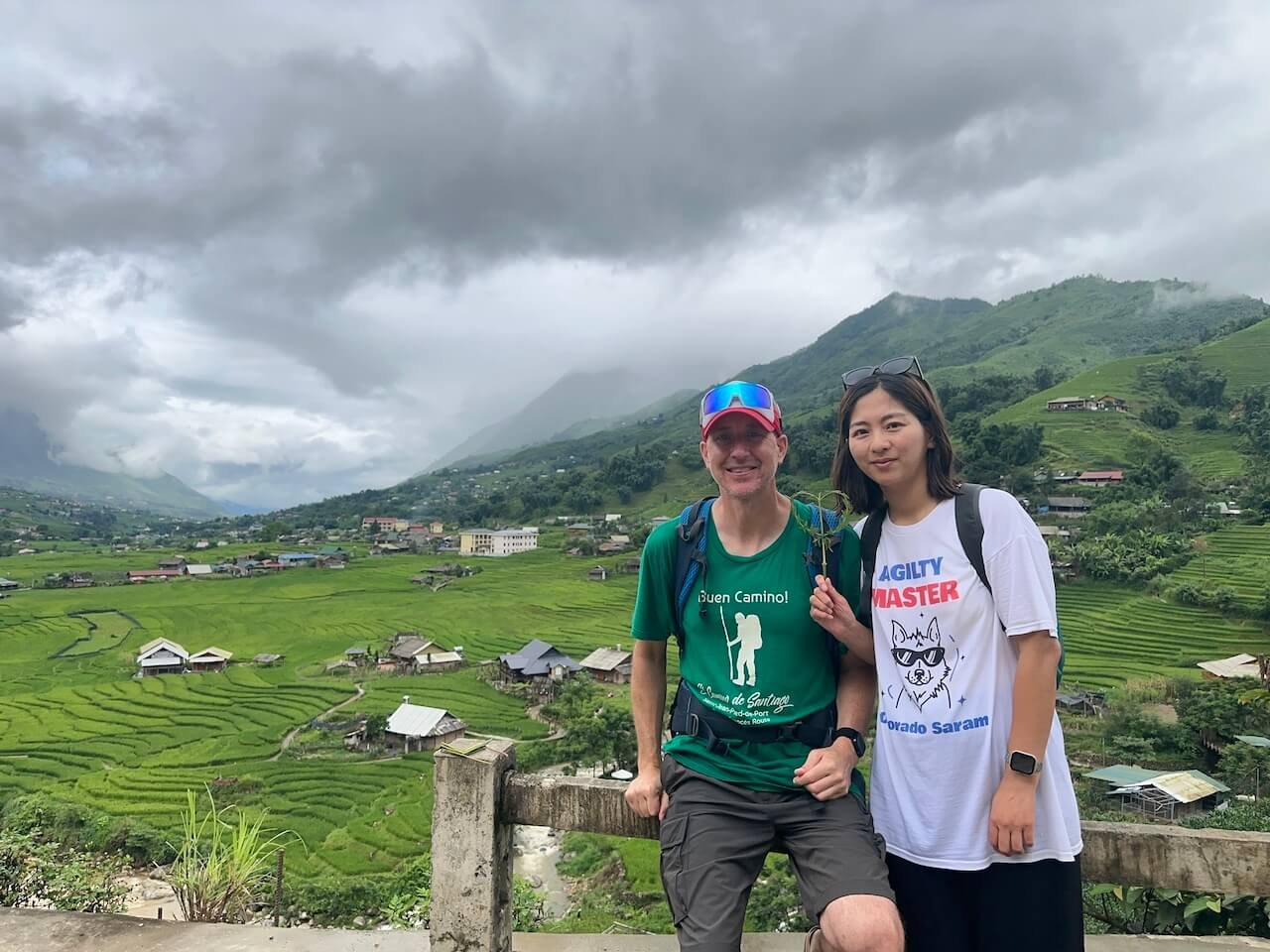 A couple trekking down a trail into Lao Chai Village in Sapa, Vietnam, with lush rice fields and mountains in the background.