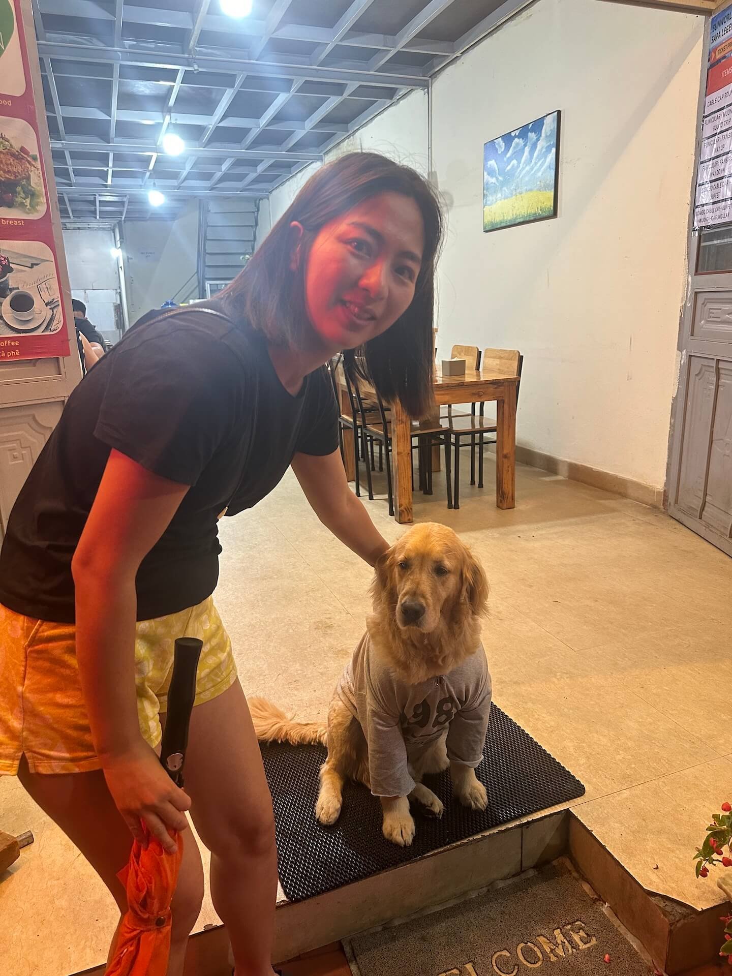 A woman smiling while petting a golden retriever wearing a gray shirt at the entrance of a restaurant in Sapa, Vietnam.