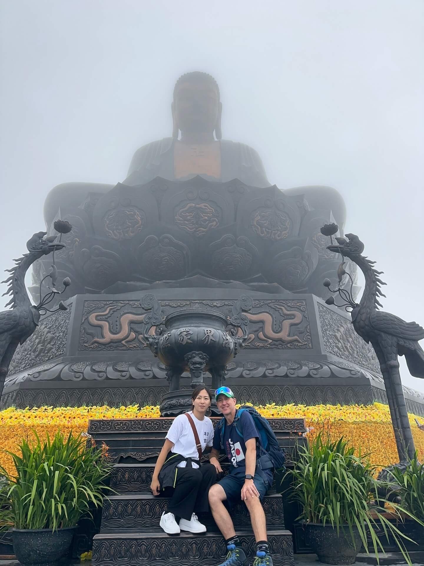 A couple sits on the steps in front of the massive Buddha statue at Fansipan Peak, with mist and golden flower decorations surrounding the base.