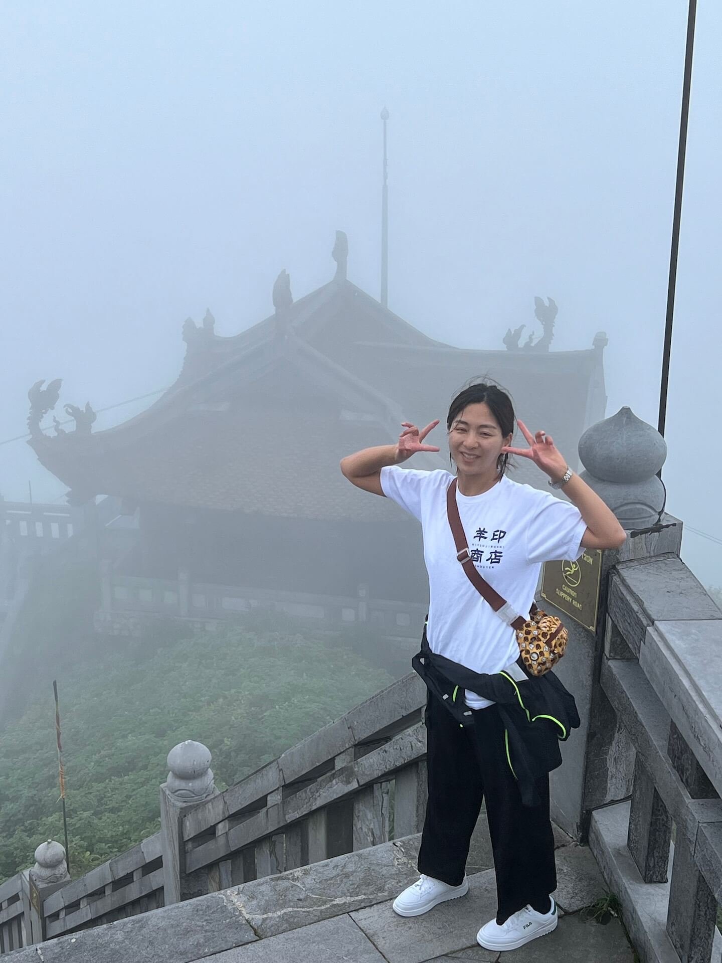 A woman poses with a peace sign on the stone stairs at Fansipan Peak, with a mist-covered temple in the background.