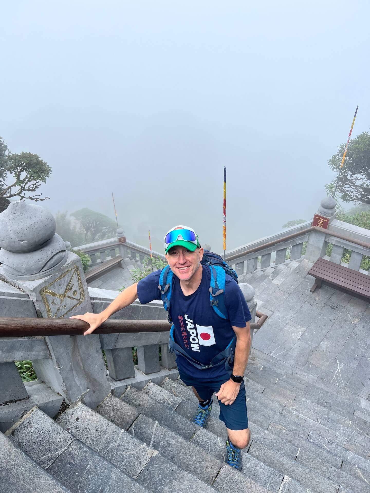 A man wearing a blue backpack and a "Japow" shirt climbs the stone stairs at Fansipan Peak, surrounded by misty weather.