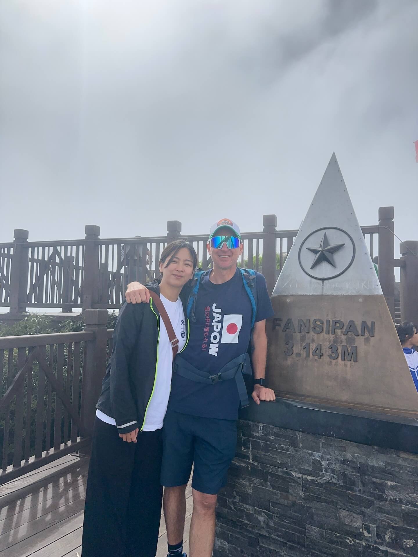 A couple posing at the summit of Fansipan Peak in Vietnam, standing in front of the elevation sign. The misty mountain backdrop adds to the dramatic atmosphere of the highest peak in Indochina.