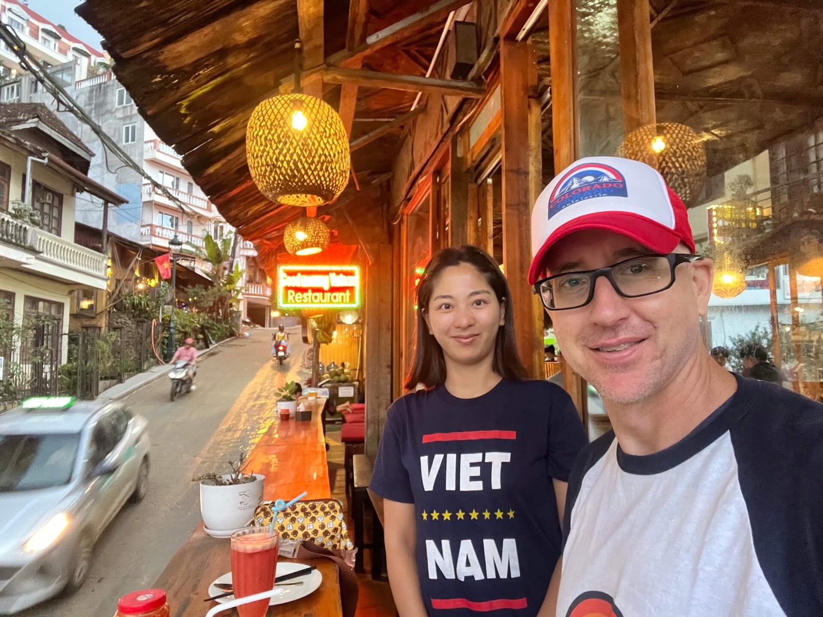 A couple enjoying dinner on the front balcony of Good Morning Vietnam restaurant in Sapa, Vietnam. The street is lively with motorbikes and cars passing by, and the restaurant's warm lighting creates a cozy atmosphere.