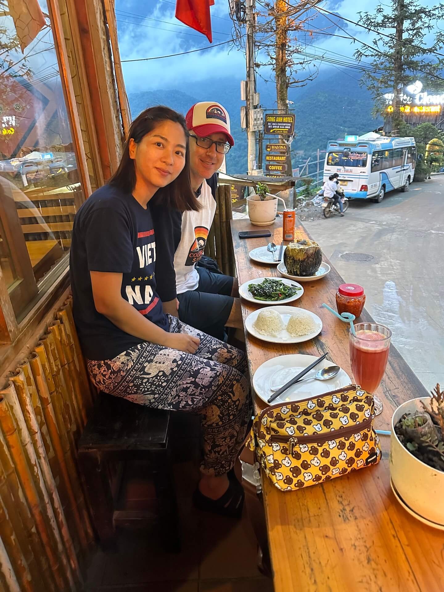 A couple dining on the balcony of Good Morning Vietnam restaurant in Sa Pa, Vietnam, with a scenic mountain view in the background. The table is set with traditional Vietnamese dishes, rice, and a fresh juice.