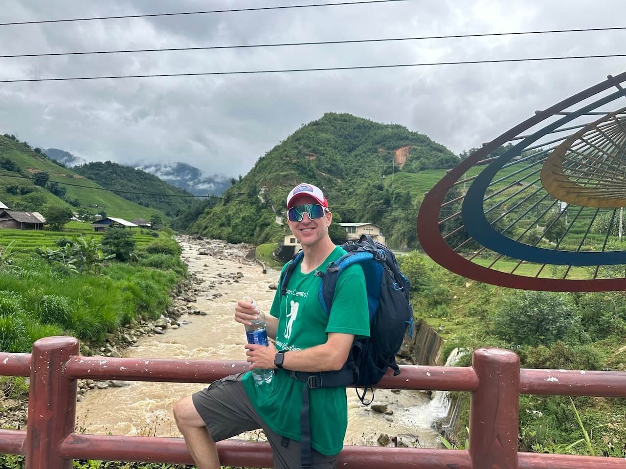 A traveler posing on a bridge over the Muong Hoa River in Sapa, Vietnam, with the scenic river and terraced rice fields in the background.