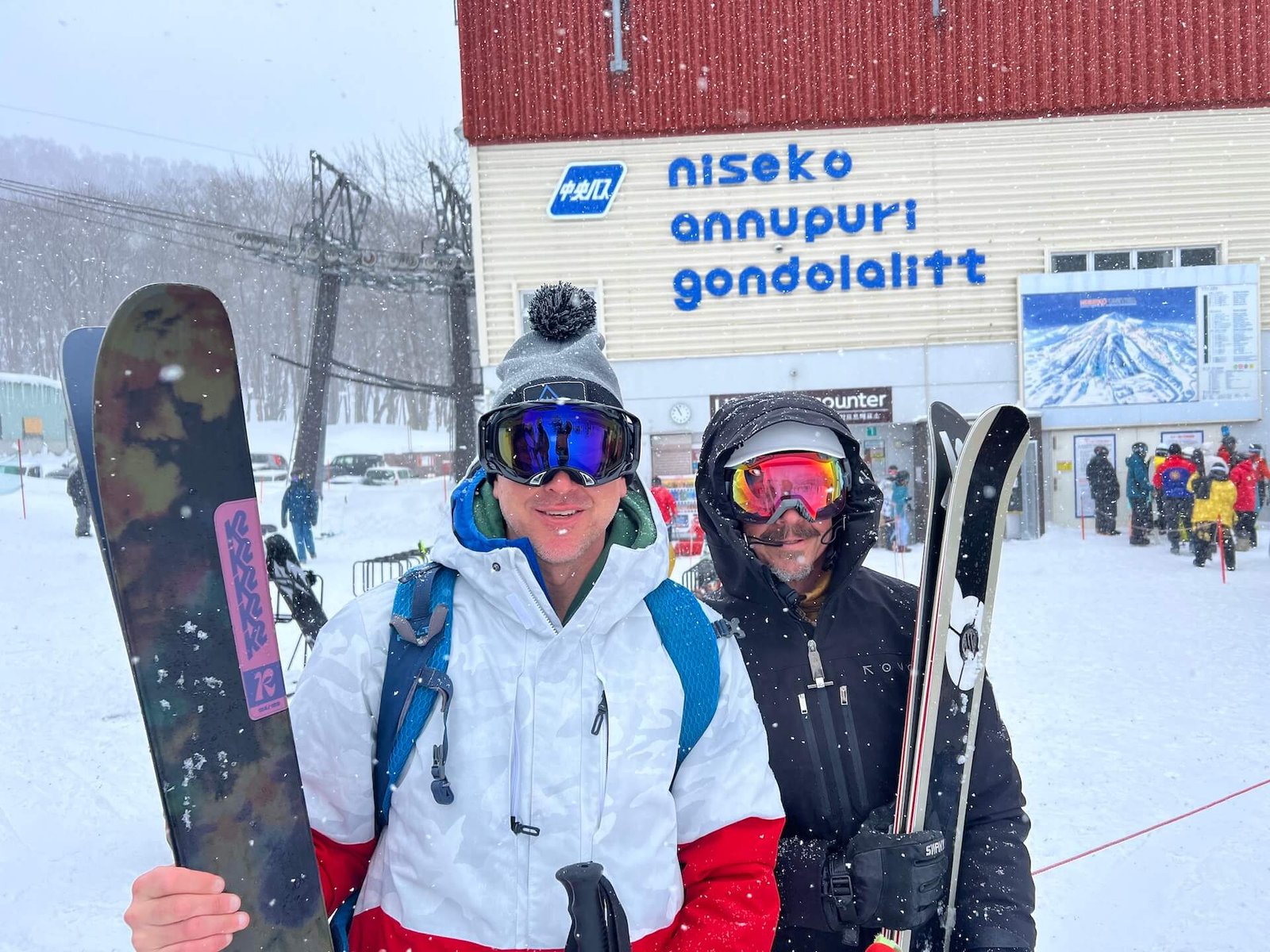 Two skiers standing at the base of Niseko Annupuri Gondola in Hokkaido, Japan, during a heavy snowfall. Dressed in ski gear and holding their skis, they are ready to experience the famous Japow powder conditions. The Niseko Annupuri gondola station and other skiers are visible in the background.