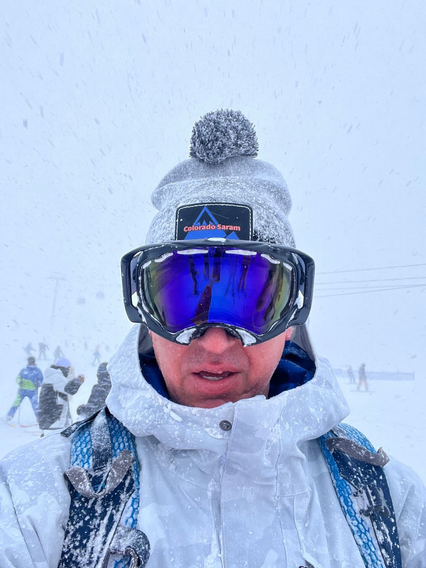 Skier taking a selfie at the base of Niseko on a heavy powder day in Hokkaido, Japan. Dressed in a white ski jacket, mirrored goggles, and a beanie with a Colorado Saram patch, they are covered in fresh snow as skiers and gondolas fade into the snowy background.