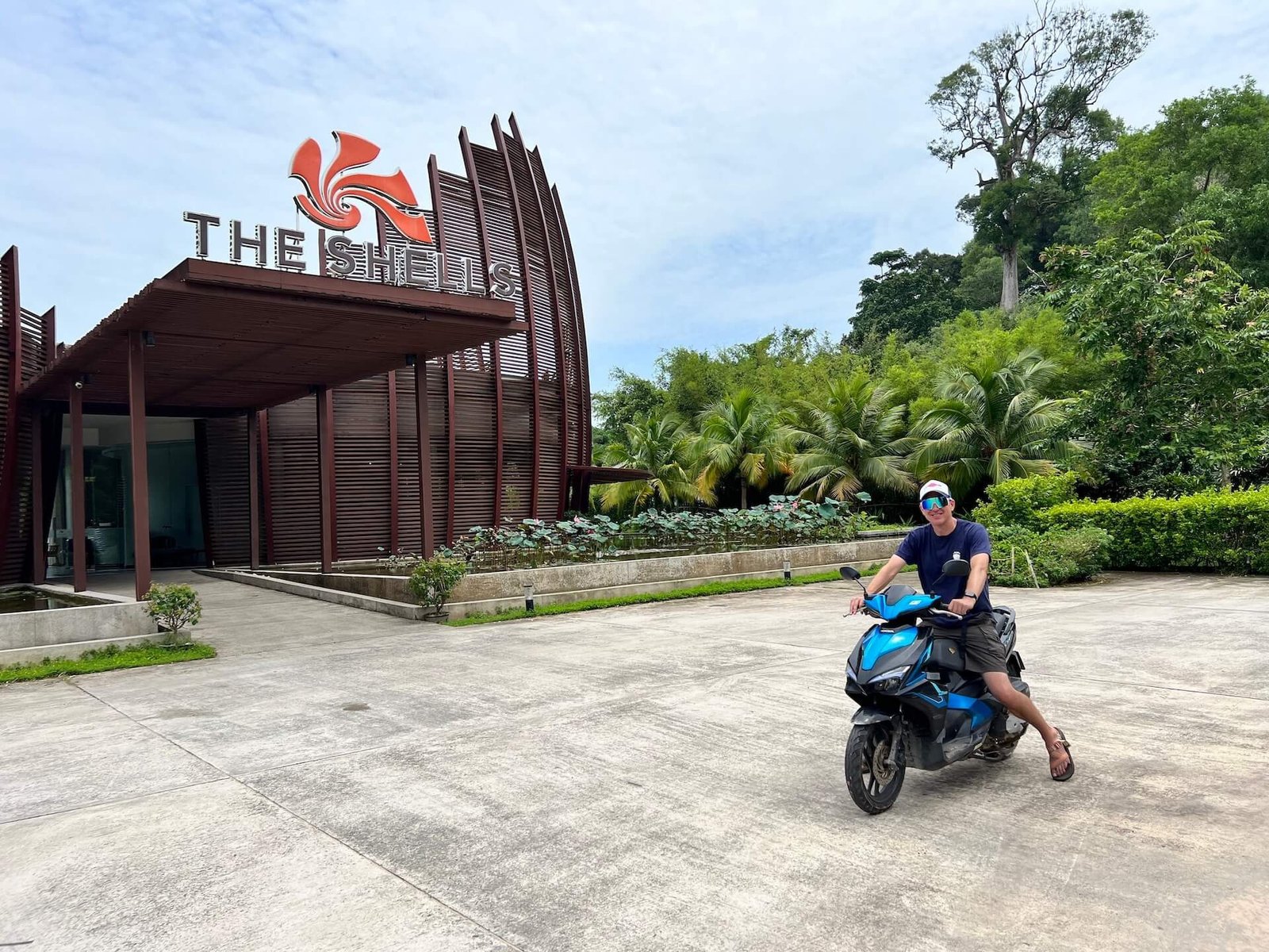 A man wearing sunglasses and a navy shirt sitting on a blue motorbike in front of The Shells Resort on Phu Quoc Island, Vietnam, surrounded by lush greenery.