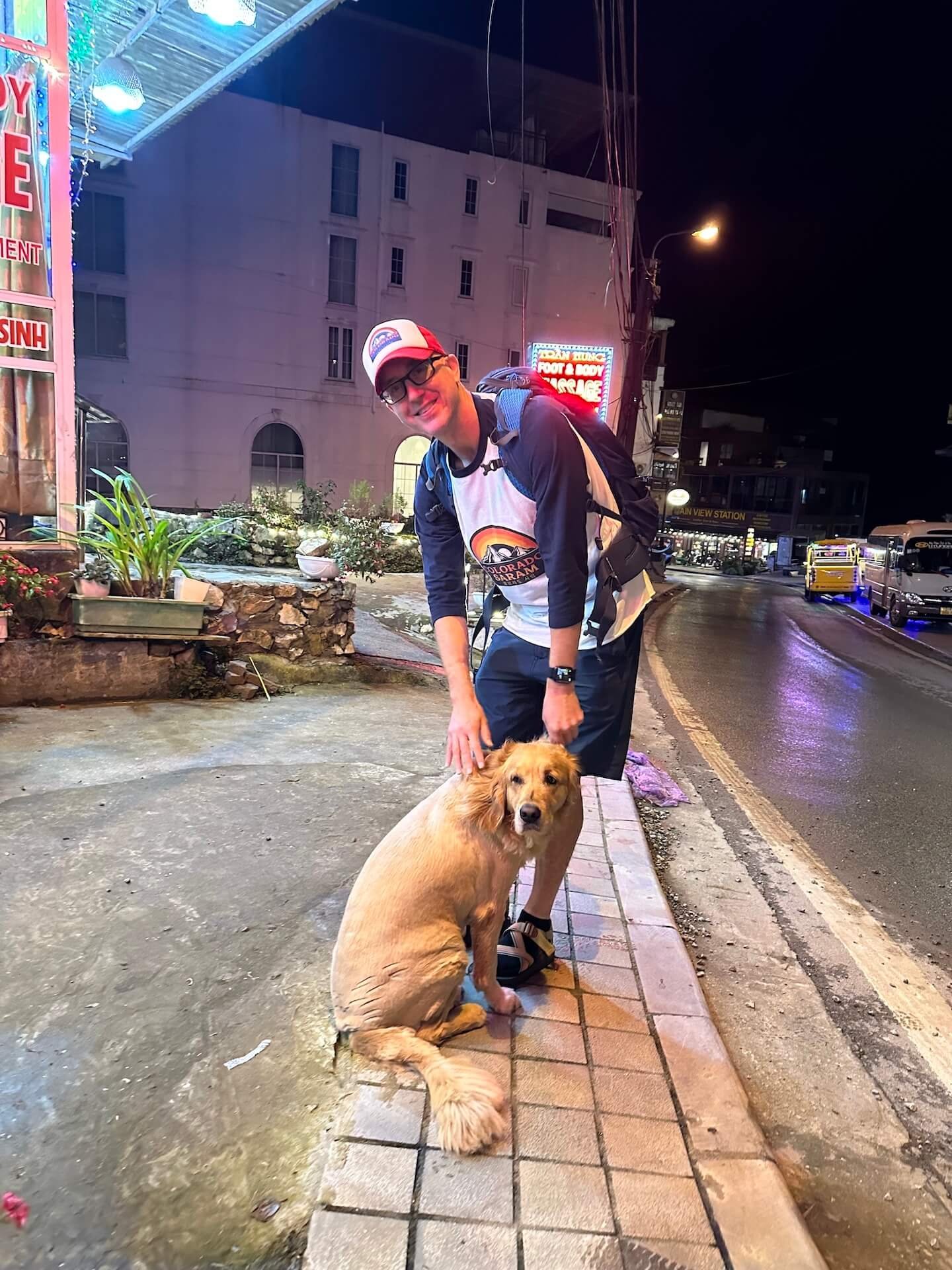  A friendly golden retriever with a short haircut sitting on the sidewalk in Sapa, Vietnam, while a man in a backpack and a Colorado Saram shirt pets it.