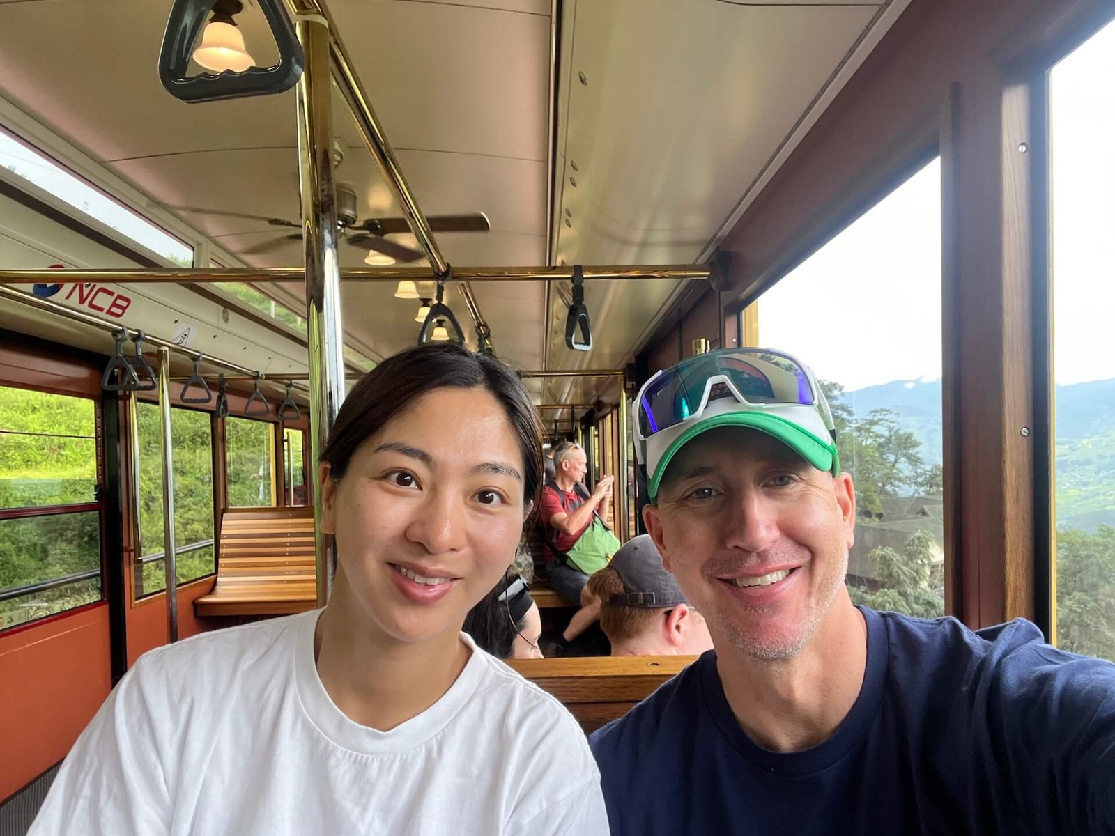 Two travelers smiling inside the Sapa monorail, which runs from Sapa Station to the Fansipan Peak cable car station, with scenic mountain views in the background.