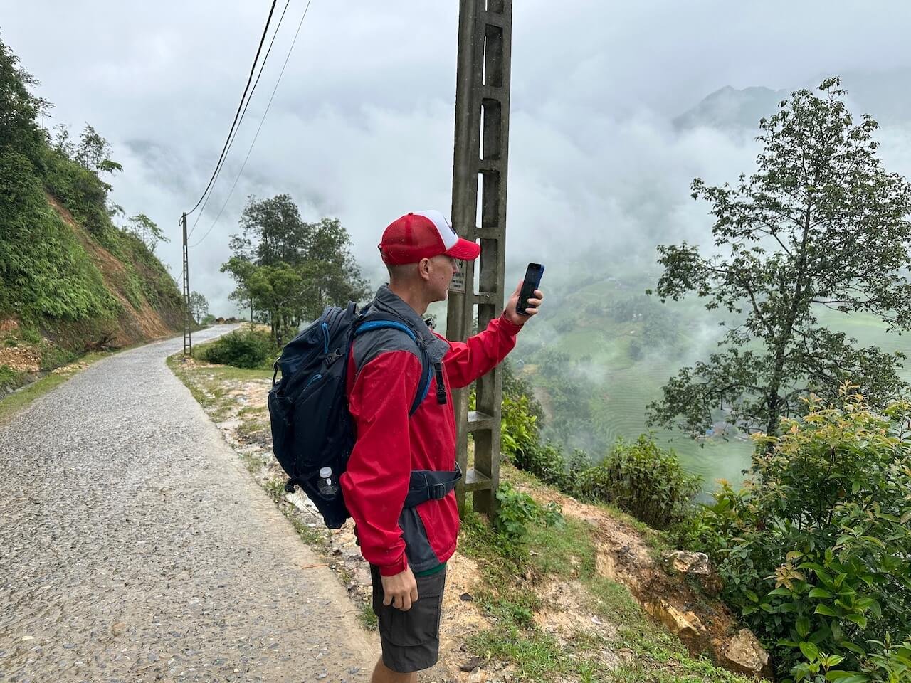 A traveler checking their phone on a paved trekking path in Sapa, Vietnam, with a winding road descending into the valley below.