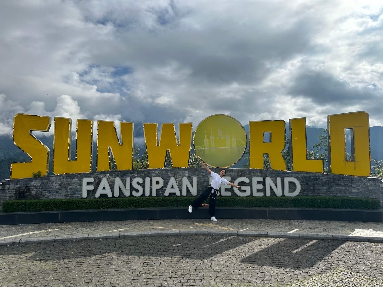 A woman posing in front of the massive Sun World Fansipan Legend sign, with misty mountains and clouds in the background at the entrance to the cable car station in Sapa, Vietnam.