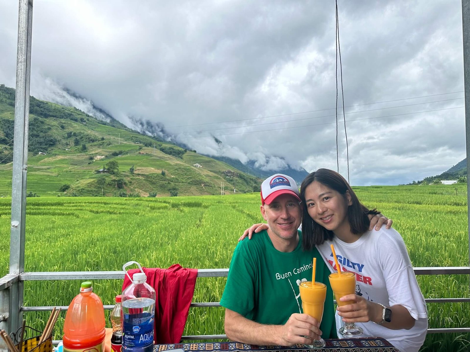 A couple enjoying mango smoothies at The Local Restaurant in Lao Chai Village, Sapa, with vibrant green rice fields and mountains in the background.