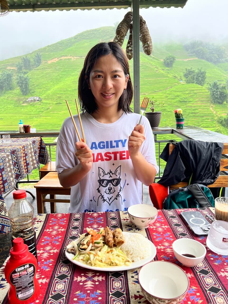 A woman sits at a table in Cúc Restaurant & Coffee, holding chopsticks and a fork, ready to enjoy a meal of rice, spring rolls, and vegetables. The open-air setting offers a stunning view of rice terraces along the trekking route from Sapa to Lao Chai.