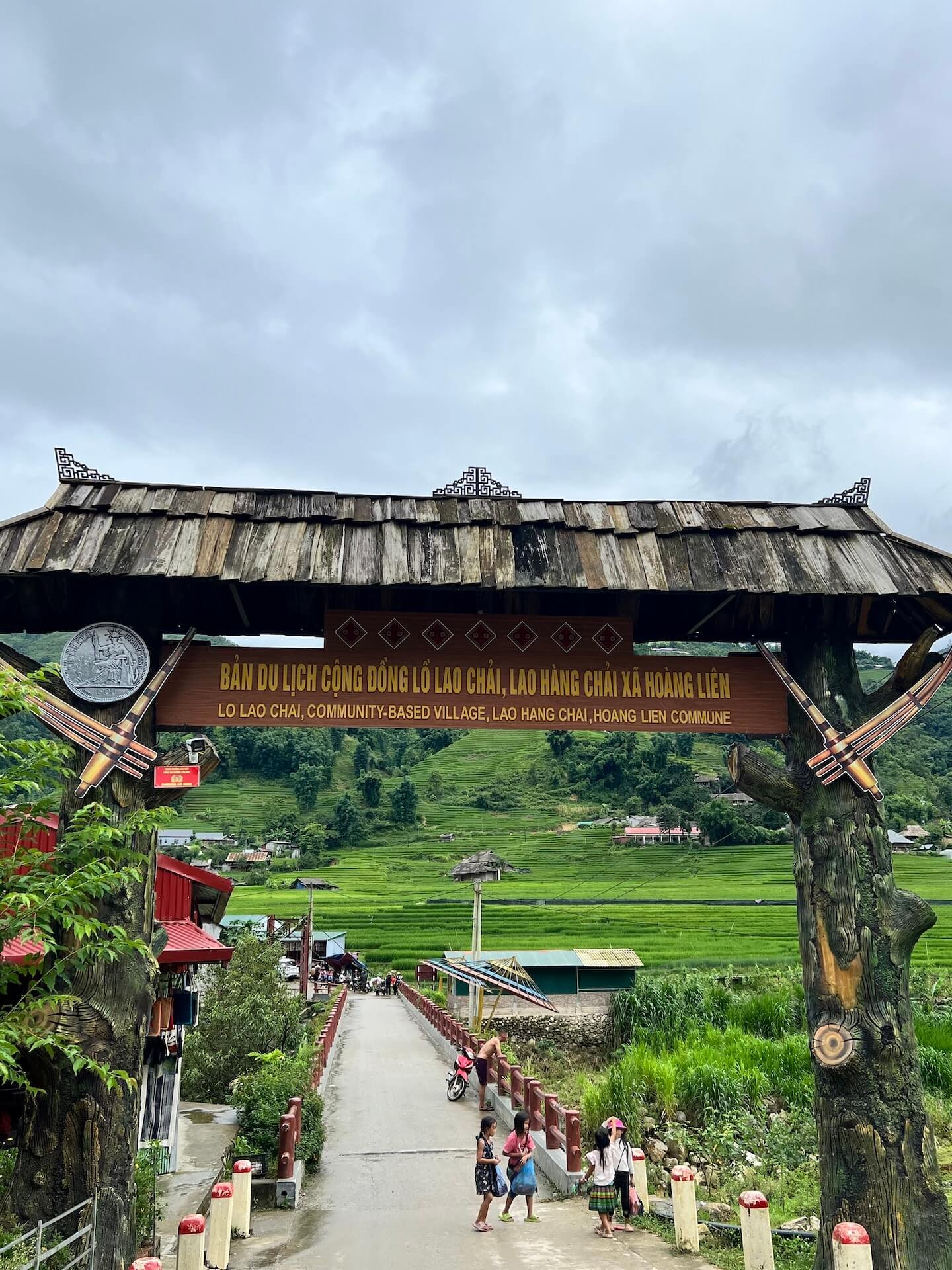 A wooden sign welcoming visitors to Lao Chai Village, a community-based village in Sapa, Vietnam, surrounded by lush greenery and mountains.