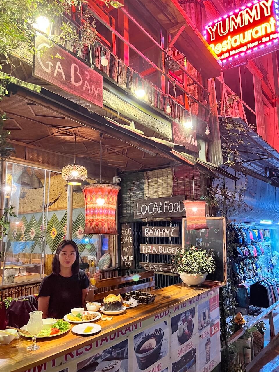 A woman sitting at an outdoor bar with traditional Vietnamese dishes at Yummy Restaurant in Sa Pa, Vietnam, with glowing lanterns and a neon sign above.