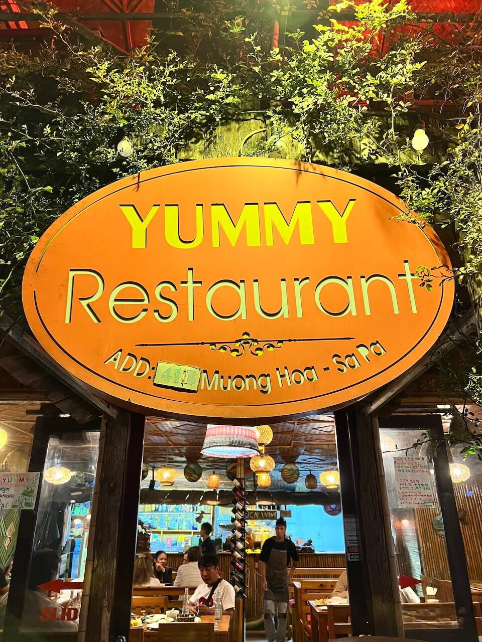 A bright orange sign reading "Yummy Restaurant" at night, with a warmly lit interior visible through the entrance in Sapa, Vietnam.