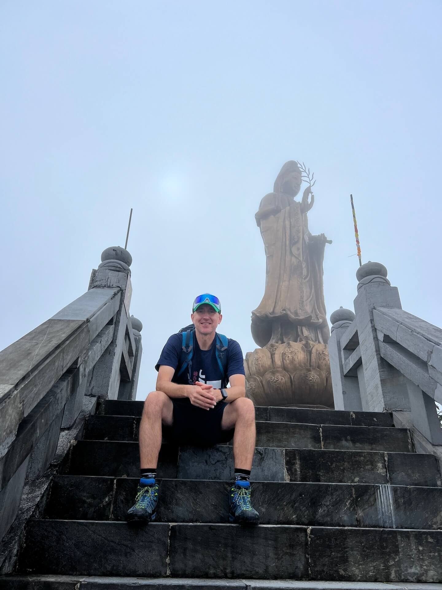Fansipan Peak Cable Car Guide: Cost, Tickets & What to Expect (2025) 13 Traveler sitting on stone stairs beneath a tall Bodhisattva statue on the Fansipan mountain temple route