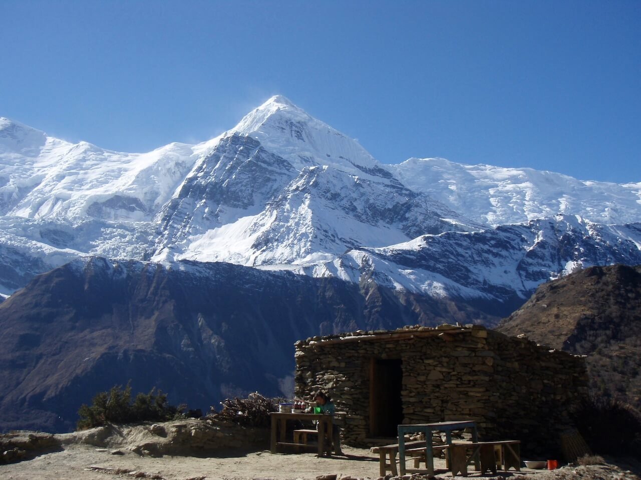Annapurna Circuit vs Everest Base Camp: How to Choose the Right Nepal Trek 7 Stone hut with wooden benches in front of dramatic snow-covered Himalayan peaks, likely near an acclimatization stop before Thorong La Pass on the Annapurna Circuit.