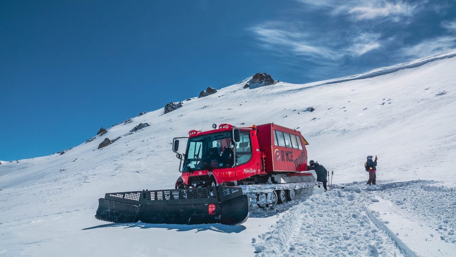 Red snowcat from Ryce Travel parked on alpine slope in Kyrgyzstan with skiers preparing to descend