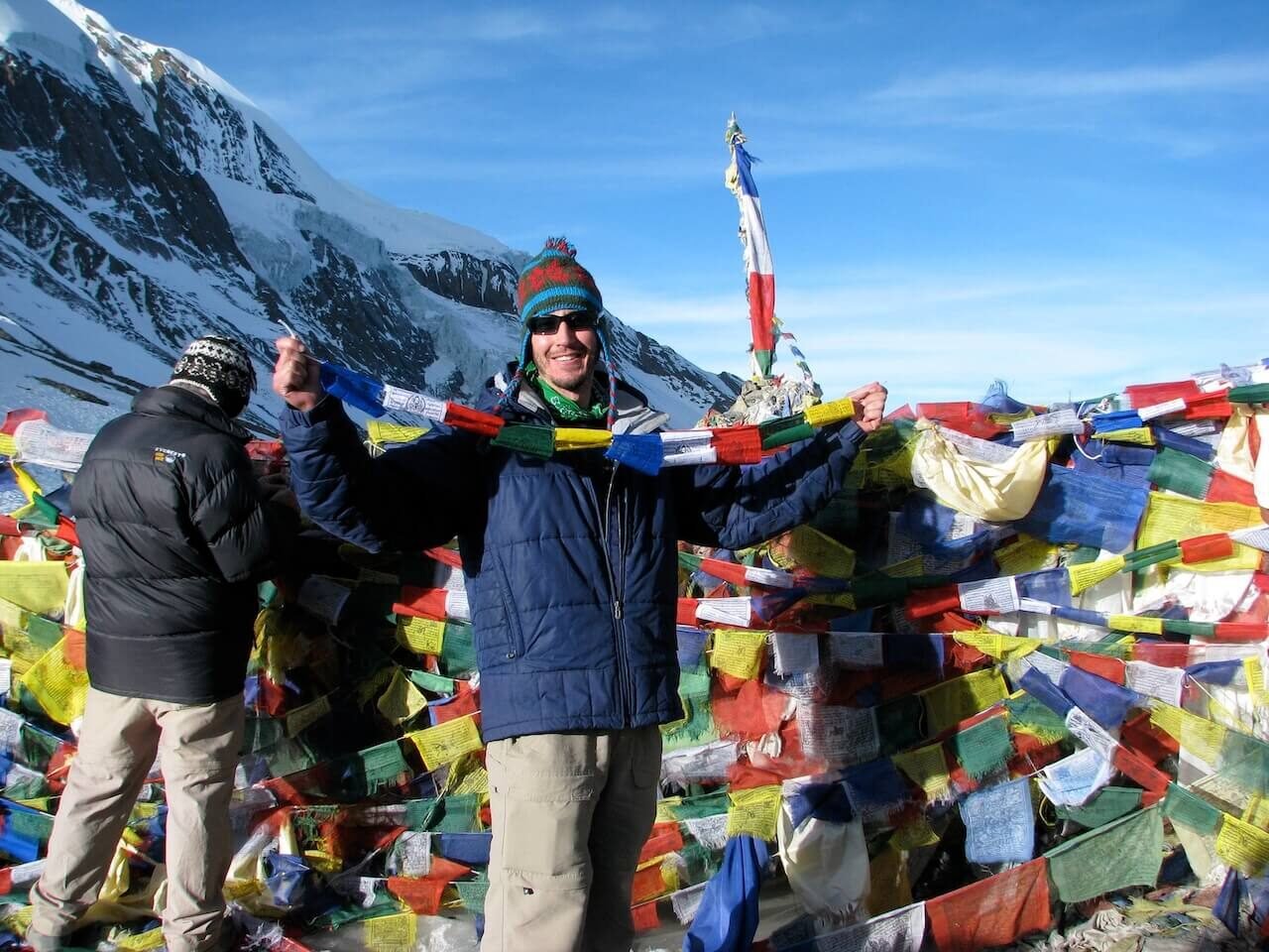 Annapurna Circuit vs Everest Base Camp: How to Choose the Right Nepal Trek 13 Trekker smiling while holding up colorful prayer flags at the snowy summit of Thorong La Pass, surrounded by mountains and traditional Himalayan prayer offerings.