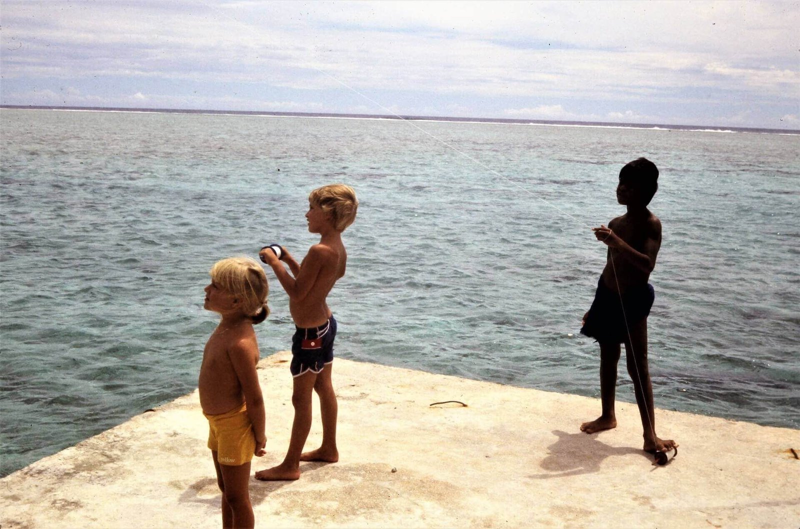 Three children flying kites from a pier over clear blue ocean water on a remote Pacific island, capturing a moment of carefree adventure.