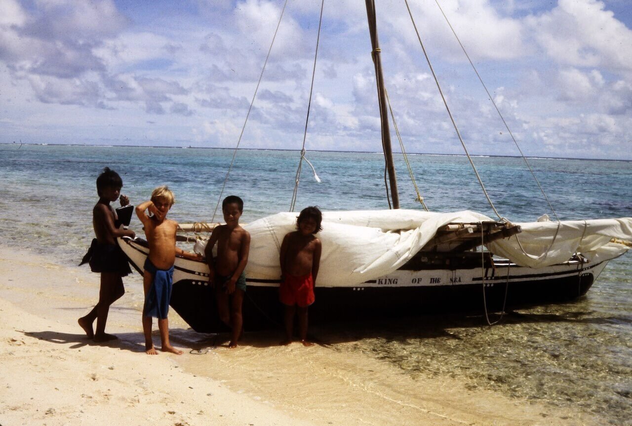John and local island friends standing beside an outrigger canoe on a beach in Micronesia, with turquoise water and a bright sky in the background.